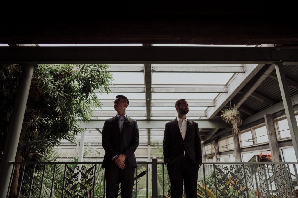 Two men in stylish suits standing thoughtfully on a balcony adorned with lush potted plants, with the overcast sky above and architectural elements of a building framing the photo