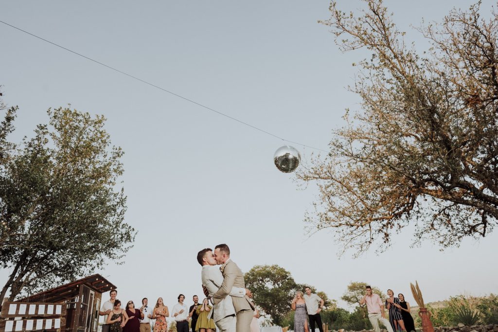 A happy moment as two grooms kiss under a disco ball hanging from a tree, with guests cheering in the background at an outdoor reception with warm evening light
