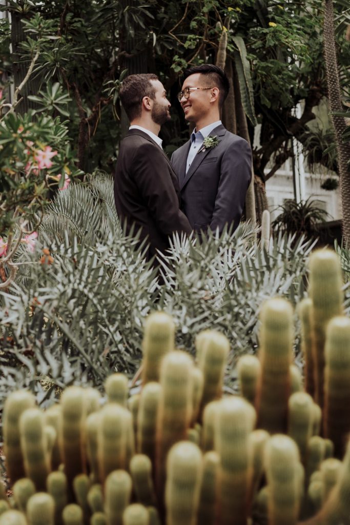 Two grooms share an intimate moment surrounded by dense, tropical greenery, with soft focus creating a romantic and secluded atmosphere in a botanical setting