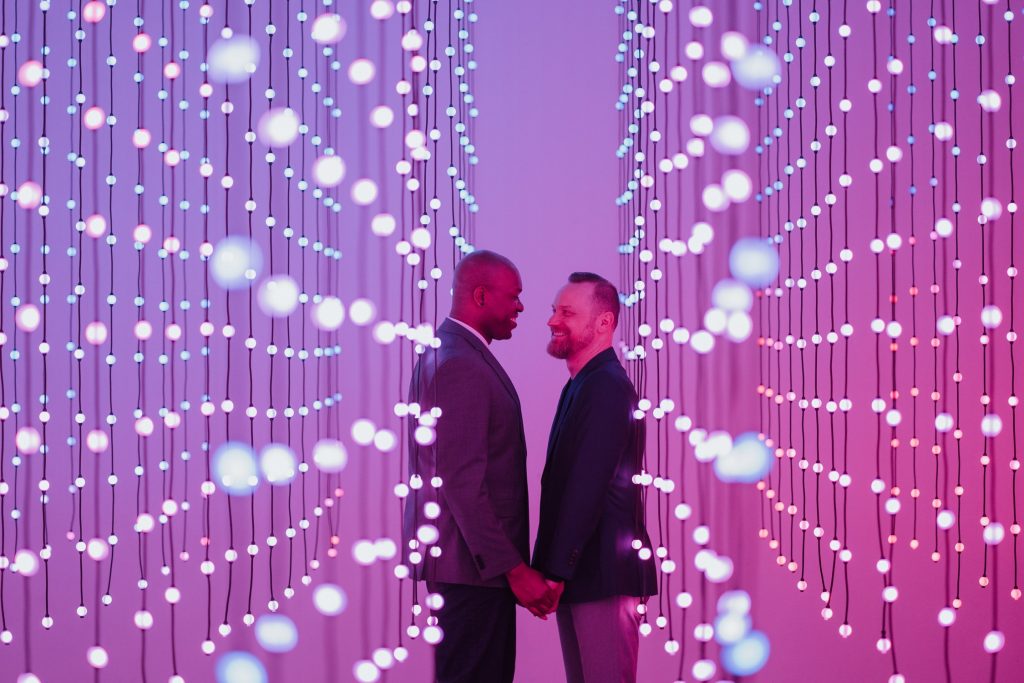 A couple stands close together, surrounded by a mesmerizing array of hanging white lights against a purple backdrop, creating an intimate and futuristic ambiance