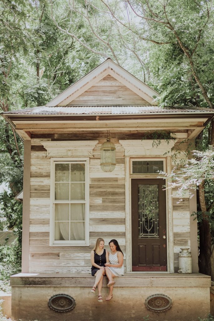 Two women sitting cozily together on the steps of a quaint, rustic wooden house, surrounded by lush trees, sharing a quiet, affectionate moment