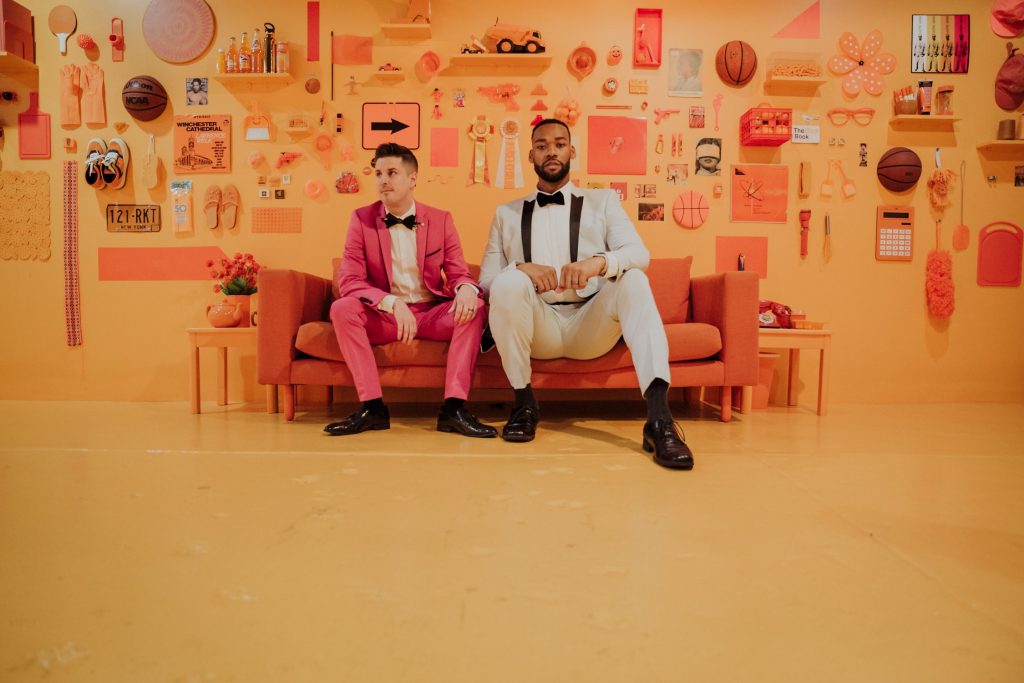 A stylish couple seated on a vibrant orange sofa in a monochromatic orange room filled with various objects, giving an artistic and contemporary vibe