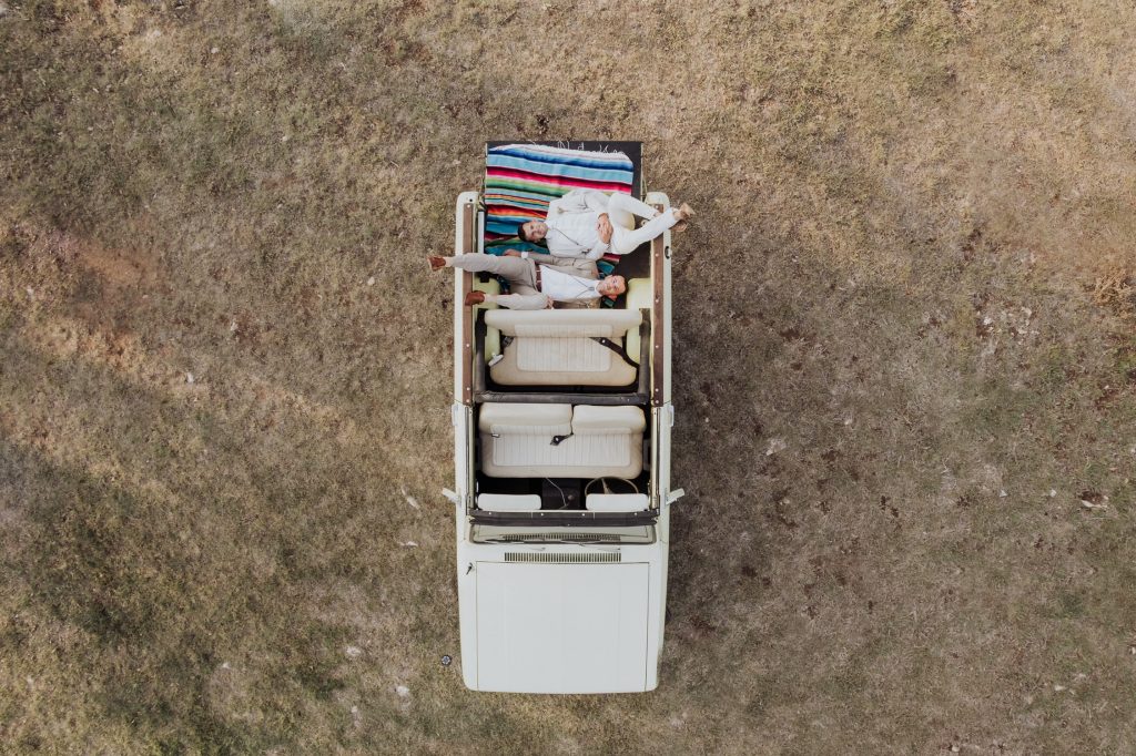 An aerial view of a couple lying on a bed placed in the back of an open vintage van, surrounded by an expansive field, depicting a whimsical and adventurous moment