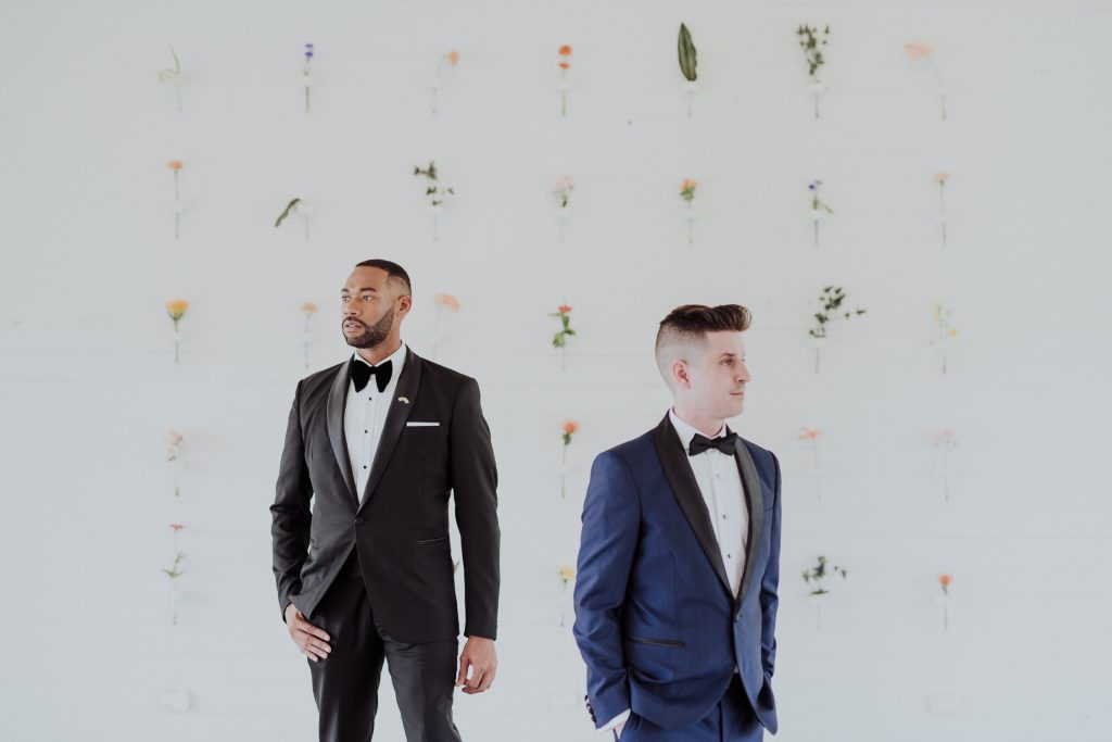 Two grooms in elegant attire stand apart, facing away from the camera against a minimalist white wall with delicate flowers, creating a serene and artistic composition