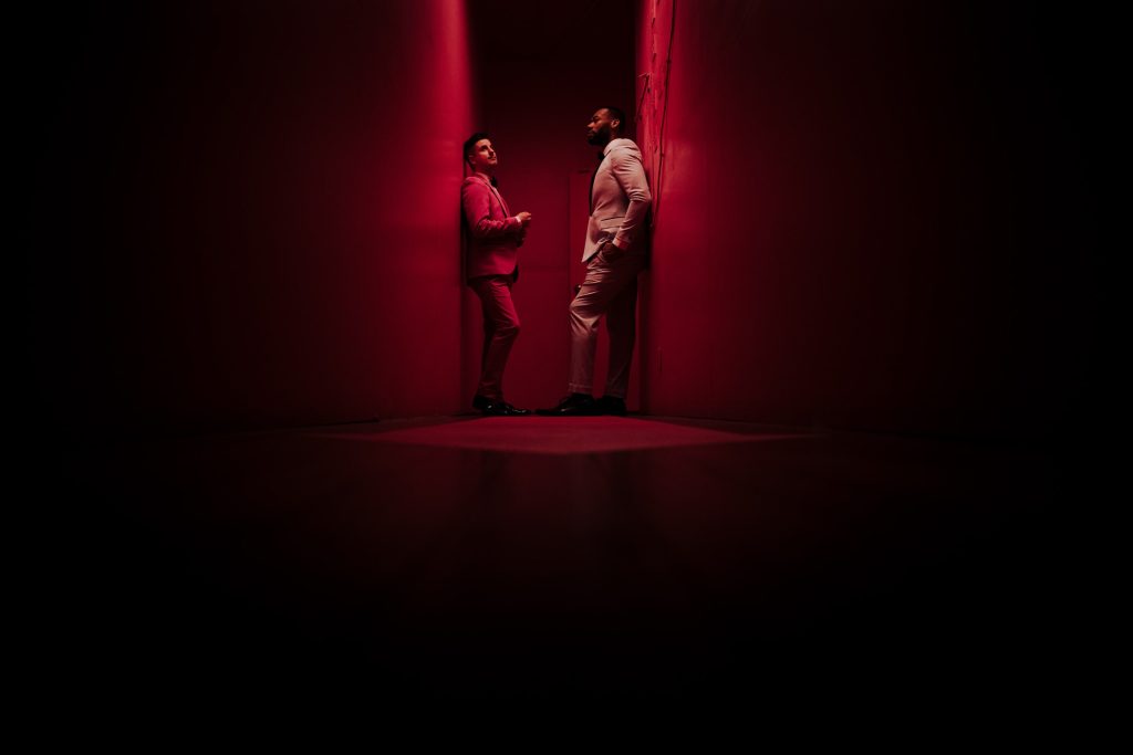 Two men in suits stand in a narrow red-lit corridor facing each other, creating a dramatic and intimate atmosphere, with one leaning casually against the wall