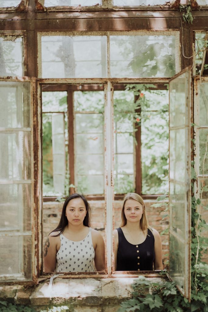 Two women stand stoically behind an aged window frame in a dilapidated greenhouse, surrounded by overgrown foliage, with a serene expression on their faces