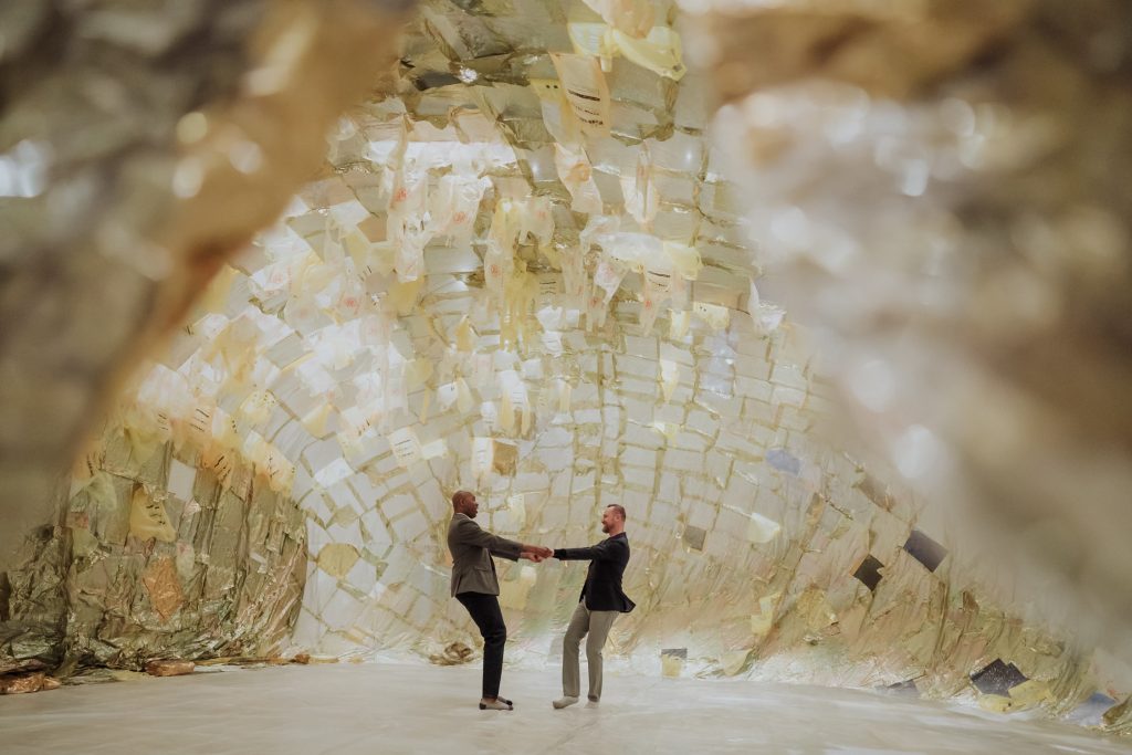 A couple holding hands stands in the middle of an art installation resembling a golden, reflective cave made from crumpled sheets of paper, giving an immersive, otherworldly feel