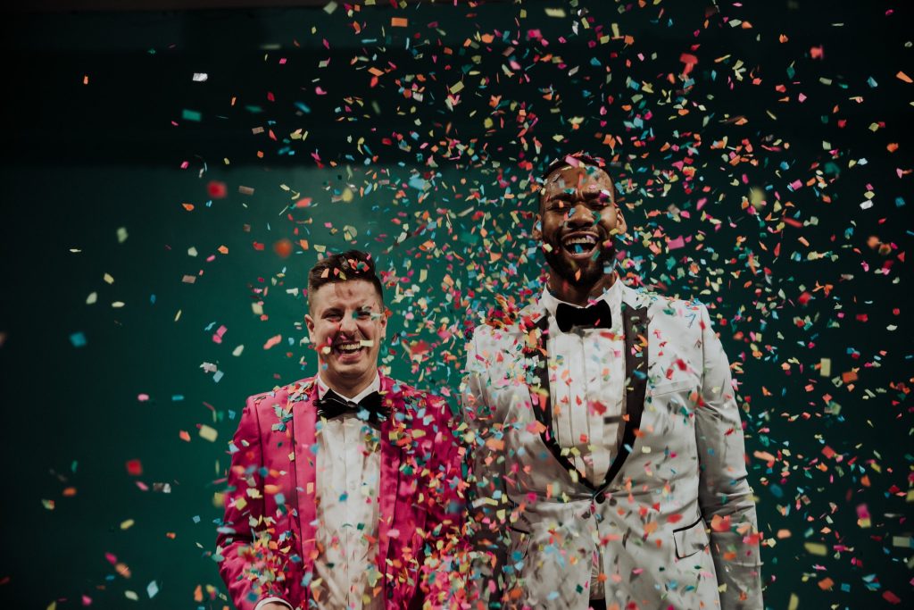 Two men in vibrant suits are covered in a shower of multicolored confetti, their joyful expressions lit by a dark teal background, celebrating a joyful moment