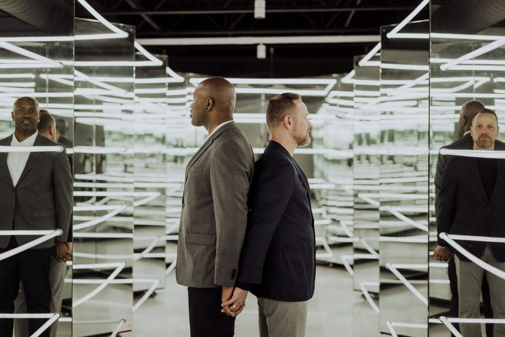 Two men in suits stand back to back in a mirrored room filled with fluorescent lights, creating an infinite reflection effect that emphasizes their contemplative stances