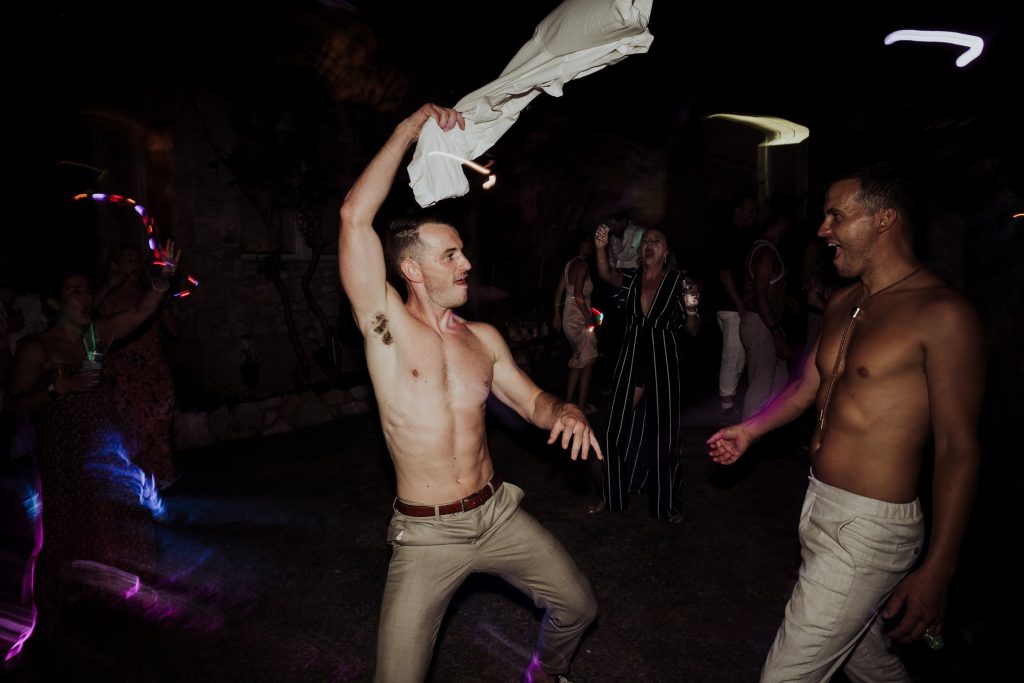 A joyful, shirtless man dances energetically at a night party, with a white shirt in hand above his head, and guests around him illuminated by streaks of colorful ligh