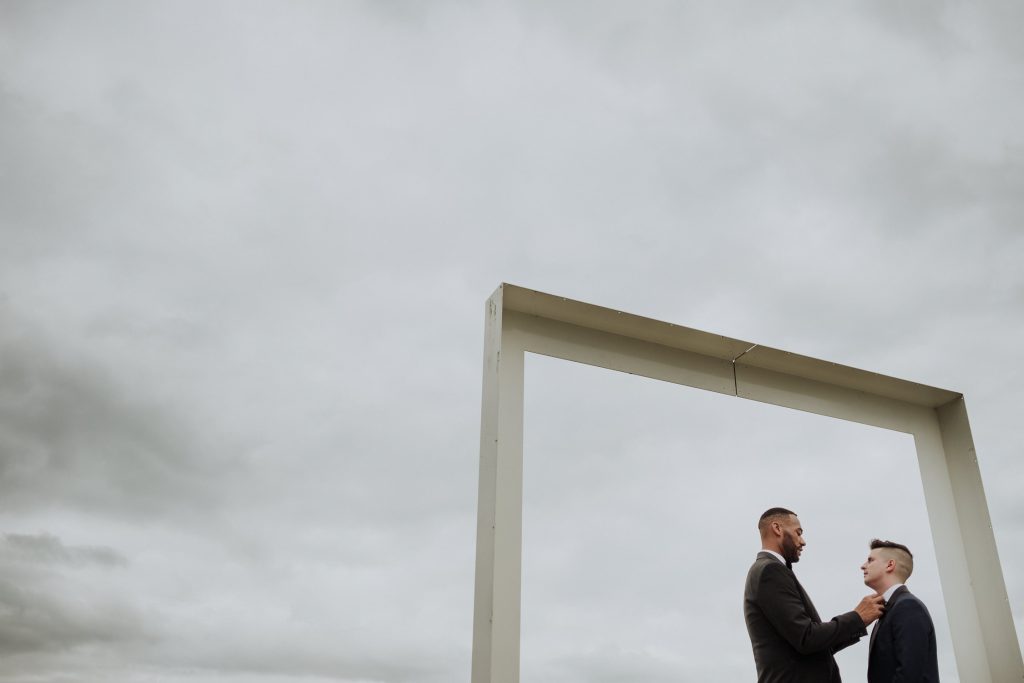 Two grooms stand under a minimalist square arch against a cloudy sky, holding each other and sharing a tender moment, symbolizing their commitment and love