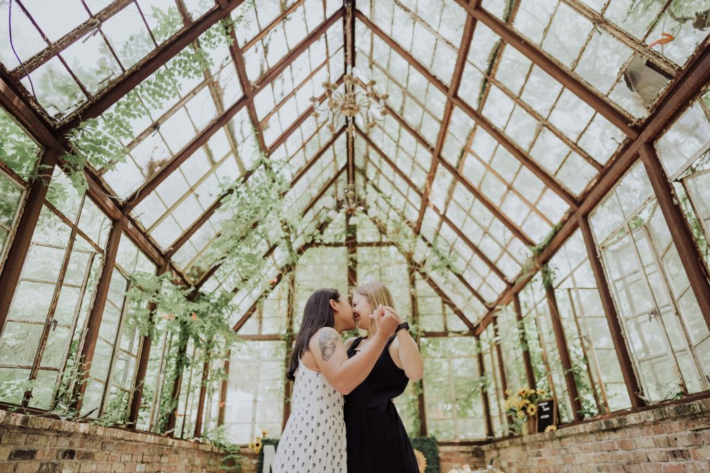 A couple kisses inside a rustic greenhouse, with sunlight filtering through the glass roof covered in green vines, creating a romantic and serene ambiance