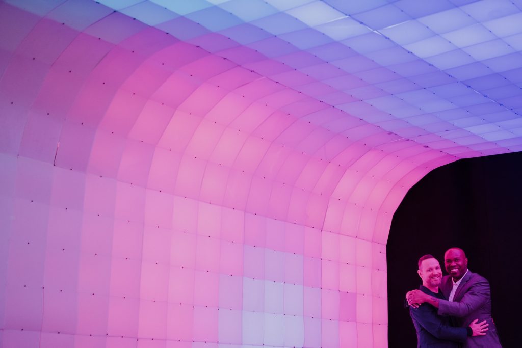 Two men embrace under a softly lit arched structure that transitions from pink to purple, their smiles suggesting a warm and heartfelt connection