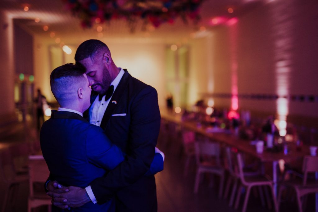 Two men in formal suits sharing an intimate moment in a dimly lit room with ambient pink and blue lighting. The background shows a blurred banquet hall setting