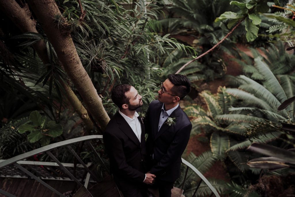 Two men in suits stand on a spiraled staircase surrounded by lush greenery and tropical plants in a greenhouse setting, sharing a loving gaze