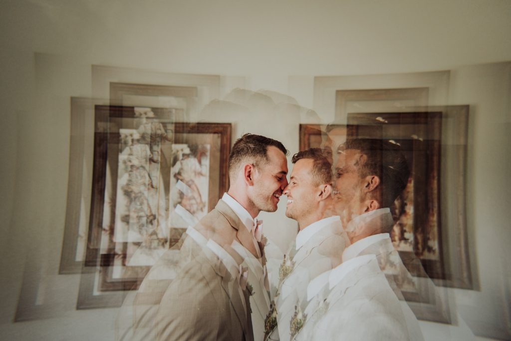 An intimate moment between two grooms in light beige suits, captured in a photograph. They are standing close, noses touching, and smiling, with the multiple exposures creating a dreamy effect of overlapping images. In the background, there's a framed piece of art, adding a touch of elegance to the tender scene