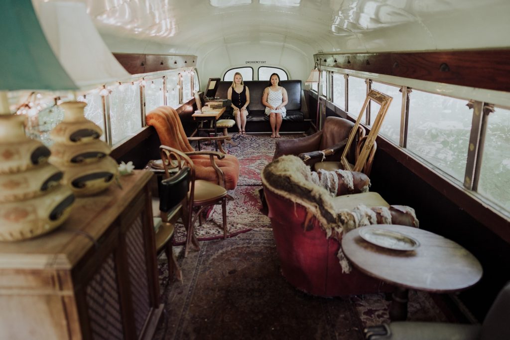 A couple, dressed in white shirts and beige pants, sitting down inside a vintage bus, converted into a cozy lounge area with eclectic furnishings
