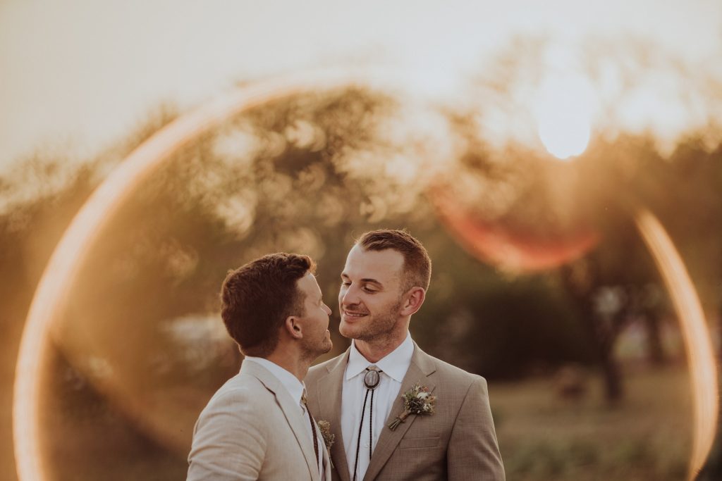 A couple in light beige suits share a kiss with a golden sun flare and an oak tree in the background