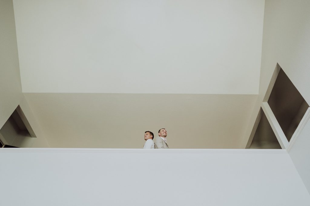 Two men dressed in light-colored suits stand together, looking down from a high vantage point in a minimalist, modern interior. The background features a plain, off-white wall and architectural elements, with a large open space in the foreground, giving a sense of tranquility and openness