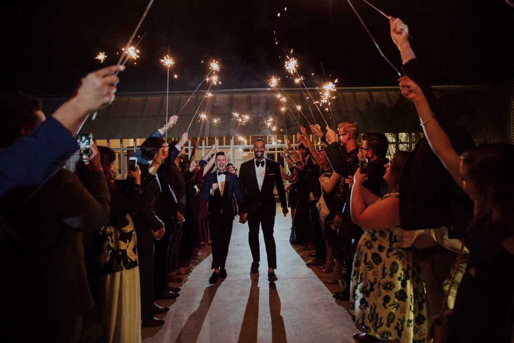 A wedding exit photo of a same-sex couple dressed in stylish suits, standing in front of a softly lit background with a hint of greenery. The lighting casts a warm, romantic glow, highlighting the affectionate embrace and the connection shared between the two individuals