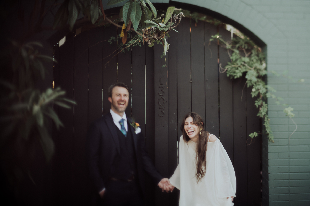 Bride and groom sharing a candid laugh in front of a charming black wooden gate adorned with greenery at Justine's Secret House in Austin, Texas