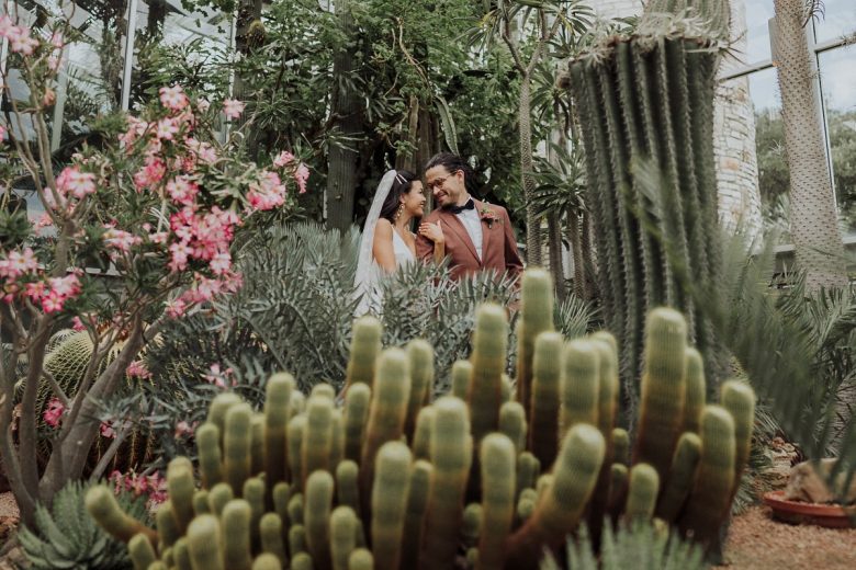 Bride and groom sharing a joyful moment surrounded by lush cacti and vibrant pink flowers in the serene setting of the Greenhouse in Driftwood, Texas