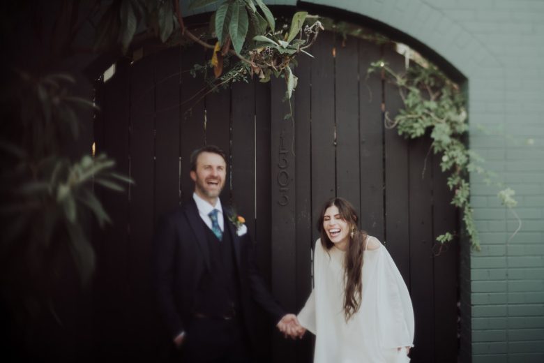 Bride and groom sharing a candid laugh in front of a charming black wooden gate adorned with greenery at Justine's Secret House in Austin, Texas
