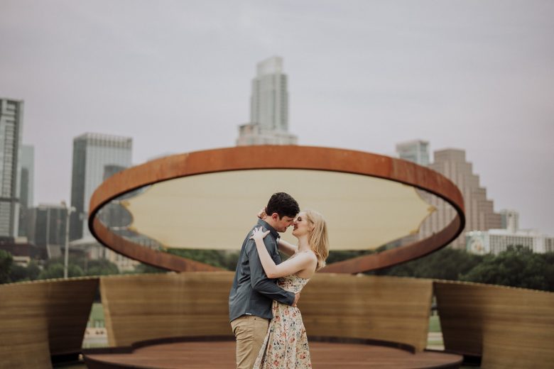 Kyra and Reno embrace beneath the circular terrace at the Long Center, with the Austin skyline behind them.