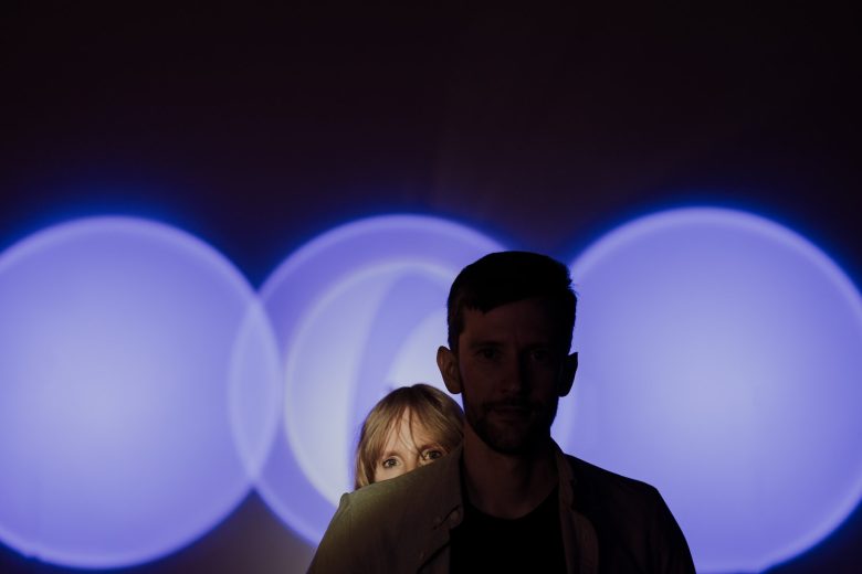 Brittany peers from behind Jeremy as blue circles of light glow around them during their Wonderspaces Austin engagement session.